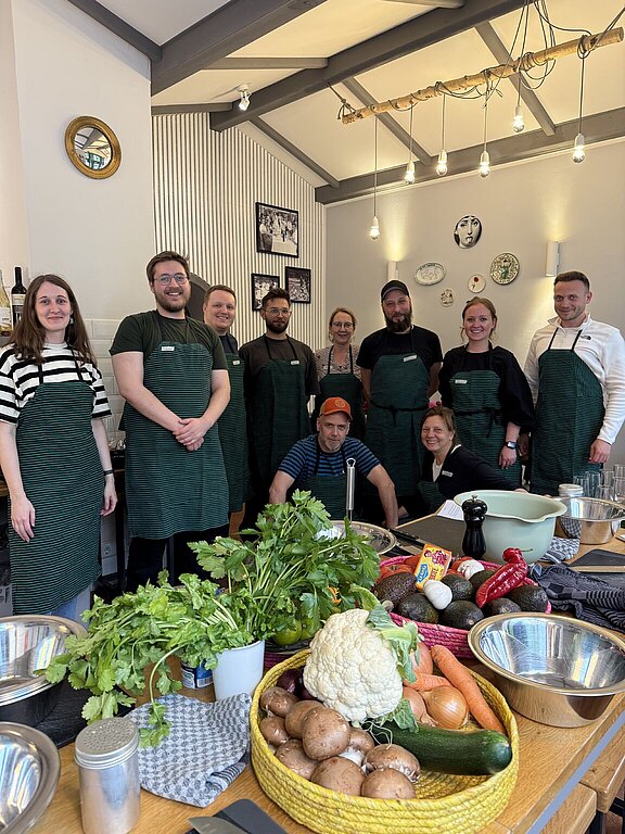 Gruppenfoto des heyday-Teams mit Kochschürzen, im Vordergrund eine mit Gemüse und Kochutensilien gefüllte Arbeitsfläche.